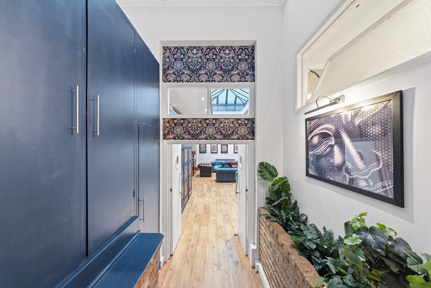 Un couloir avec une armoire bleu foncé, des carreaux muraux à motifs, un plancher en bois, un coin salon avec un canapé bleu et une œuvre d’art encadrée.
