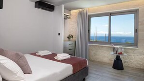 A modern bedroom with a bed, a small table with a bottle and fruit, a window with a view of the sea, and a stone wall. - Luxurious Villa Rocca Blanco 2 (Chania)