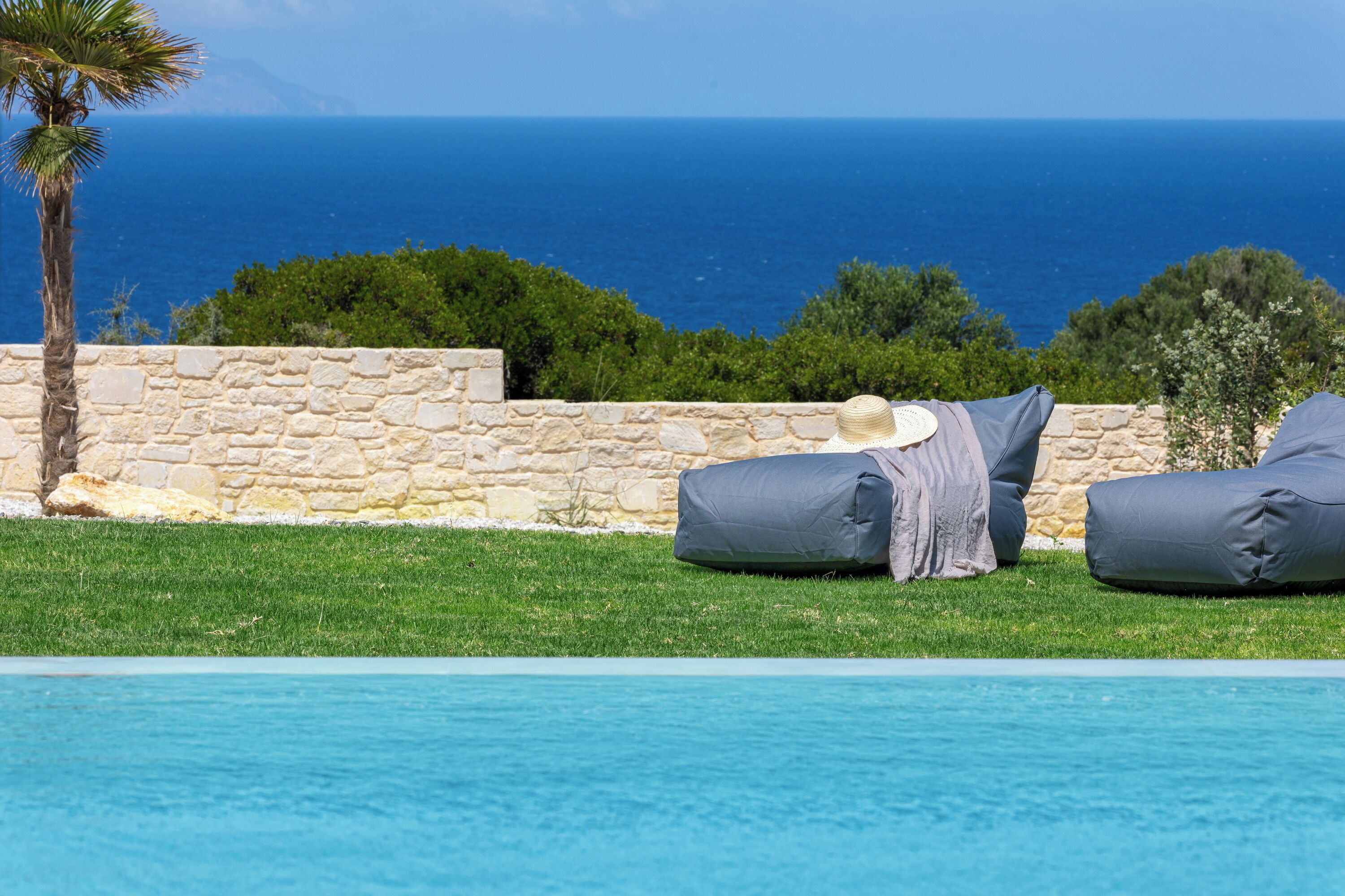 A poolside area with two grey lounge chairs, a stone wall, and a view of the sea.