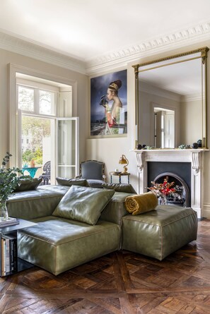 A room with a green leather sofa, a fireplace, a large mirror, and a painting on the wall. - Eccleston Square V by onefinestay (London)