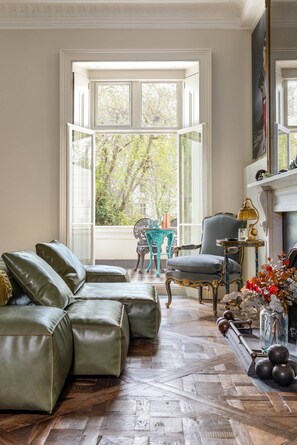 Una habitación con un ventanal grande, un sofá verde, un sillón azul, una mesita pequeña y una chimenea.