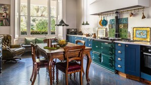 A traditional dining room with a table and chairs, a kitchen with blue cabinets and a stove, and a large window with a view of greenery. - Eccleston Square V by onefinestay (London)