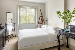 A neatly made bed with white linens, a bedside table with a vase of green plants, a large window with a view of trees, and a mirror on the wall. - Eccleston Square V by onefinestay (London)