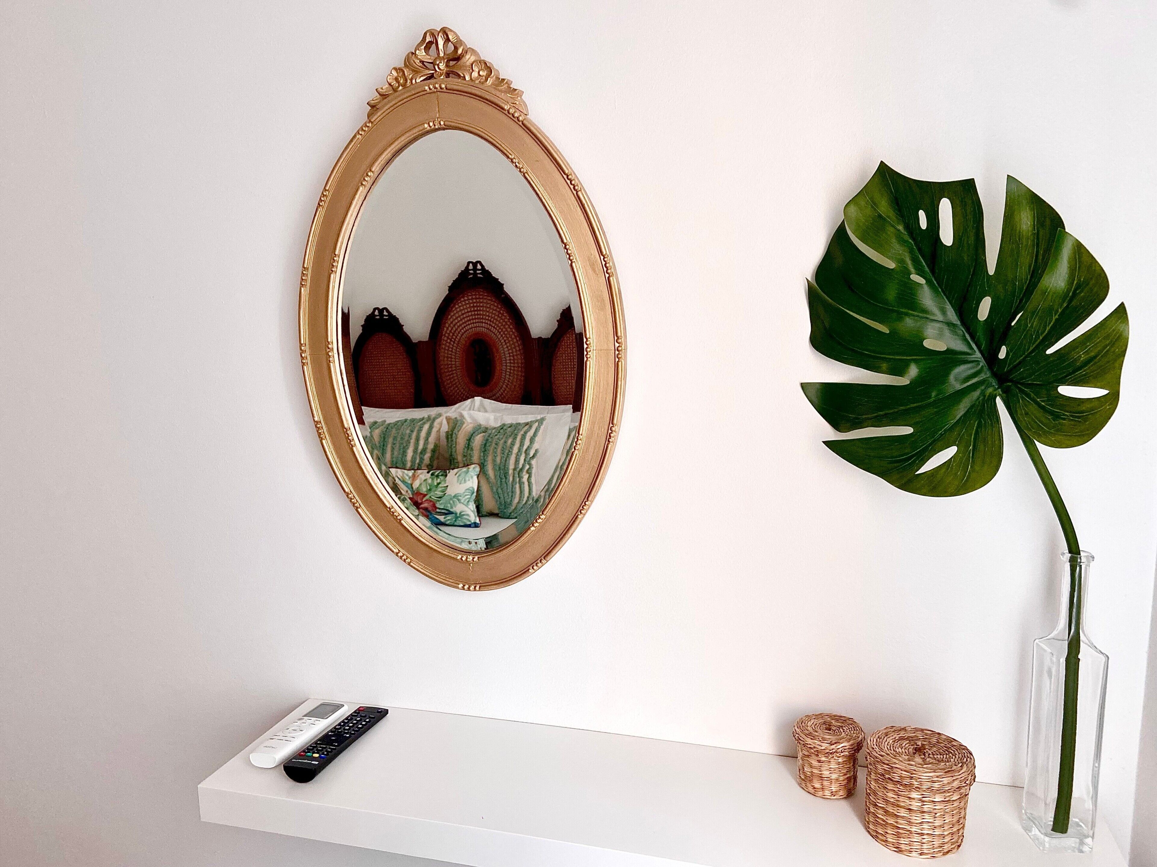 Ein weißes Regal mit einer Fernbedienung, ein großer Spiegel mit goldener Rahmung und eine Kübelpflanze mit Monstera-Blättern.