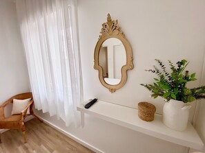 Habitació amb paret blanca, un mirall decoratiu, prestatge blanc amb comandament a distància, cistella de vimet i una planta en test.