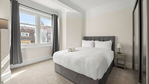 Een slaapkamer met een groot bed, een nachtkastje met een lamp, een raam met gordijnen en uitzicht op gebouwen.