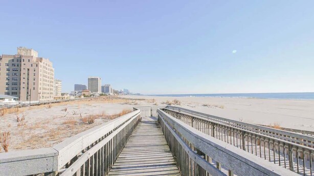 Een looppad leidt naar een strand, met gebouwen op de achtergrond.