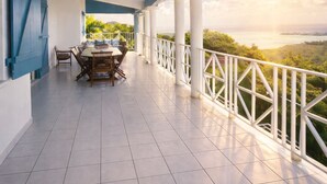 Eine überdachte Terrasse mit Esstisch und Stühlen, weißen Geländern und Blick auf eine Landschaft bei Sonnenuntergang.