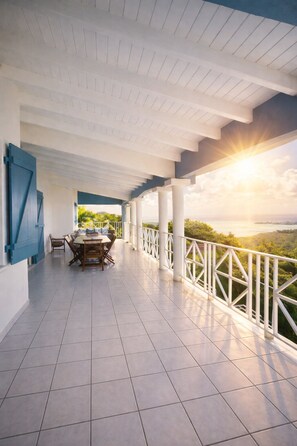 Eine überdachte Terrasse mit Esstisch und Stühlen, weißen Geländern und Blick auf eine Landschaft bei Sonnenuntergang.