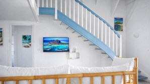Ein Schlafzimmer mit einem hölzernen Bett, weißen Wänden, einer blauen Treppe und einem Fernseher, der eine tropische Strandlandschaft zeigt.