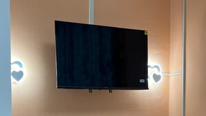 Una habitación con una televisión de pantalla plana montada en la pared, un mini refrigerador blanco y luminarias con forma de corazón.
