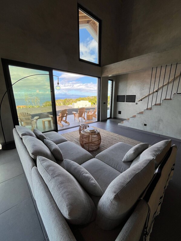 A modern living room with a large sofa, a dining area with chairs, a staircase, and a view of the ocean.