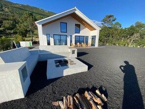A modern house with a sloped roof, large windows, and a concrete outdoor seating area. - Casa     do   Atalho (SÃO ROQUE DO PICO)