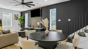 Una sala de estar moderna con una mesa redonda negra, sillas color beige, un ventilador de techo y una escalera.