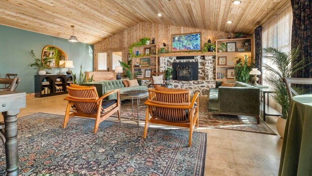 Een gezellige woonkamer met houten plafond, een open haard en een grote televisie. Er staan twee rieten fauteuils, een bank en een eettafel met stoelen.