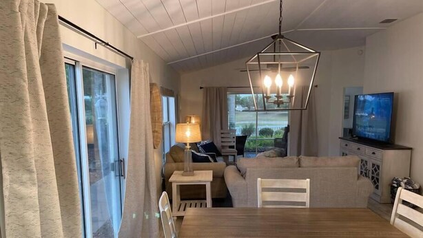 Een eethoek met een houten tafel en stoelen, een kroonluchter, een televisie en een bank op de achtergrond.