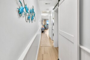 A hallway with a decorative metal wall art, a wooden floor, and a view into a room with a bed and a window.