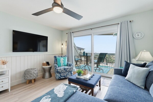 A spacious living room with a large TV, comfortable seating, a ceiling fan, and a view of the ocean through large windows.