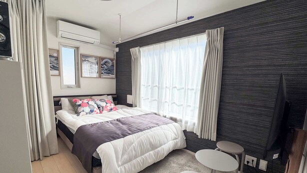 Ein modernes Hotelzimmer mit einem großen Bett, zwei kleinen Tischen, einem Flachbildfernseher und einem Fenster mit durchsichtigen Vorhängen.