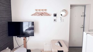 Ein Hotelzimmer mit Flachbildfernseher, einer Kleiderstange aus Holz, einem Wandspiegel, einem Thermostat, einer Fernbedienung und einer kleinen Aufbewahrungsbox.