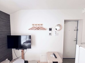A hotel room with a flat-screen TV, a wooden hanger rack, a wall-mounted mirror, a thermostat, a remote control, and a small storage box. - Triphome Shinokubo Cozy Stay (Shinjuku City)