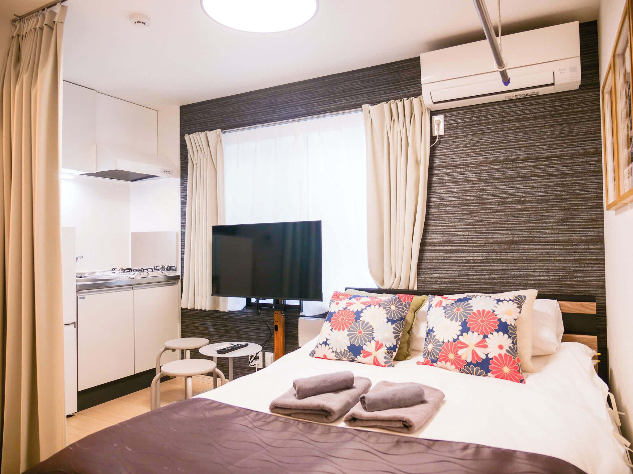 A hotel room with a bed, a TV, a small table, and a kitchenette.