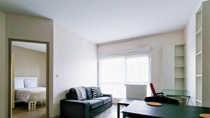 Ein modernes, helles Zimmer mit einem Bett, einer Couch, einem Esstisch und einem großen Fenster.