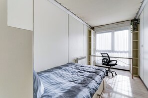 Ein Schlafzimmer mit Bett, Schreibtisch und Stuhl. Ein Fenster mit Jalousien und eine Heizung sind ebenfalls vorhanden.