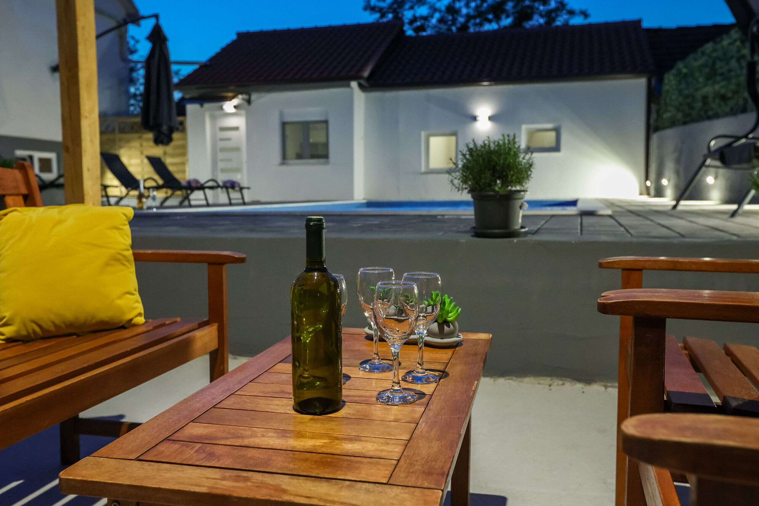 An outdoor seating area with a wooden table, a bottle, and wine glasses.