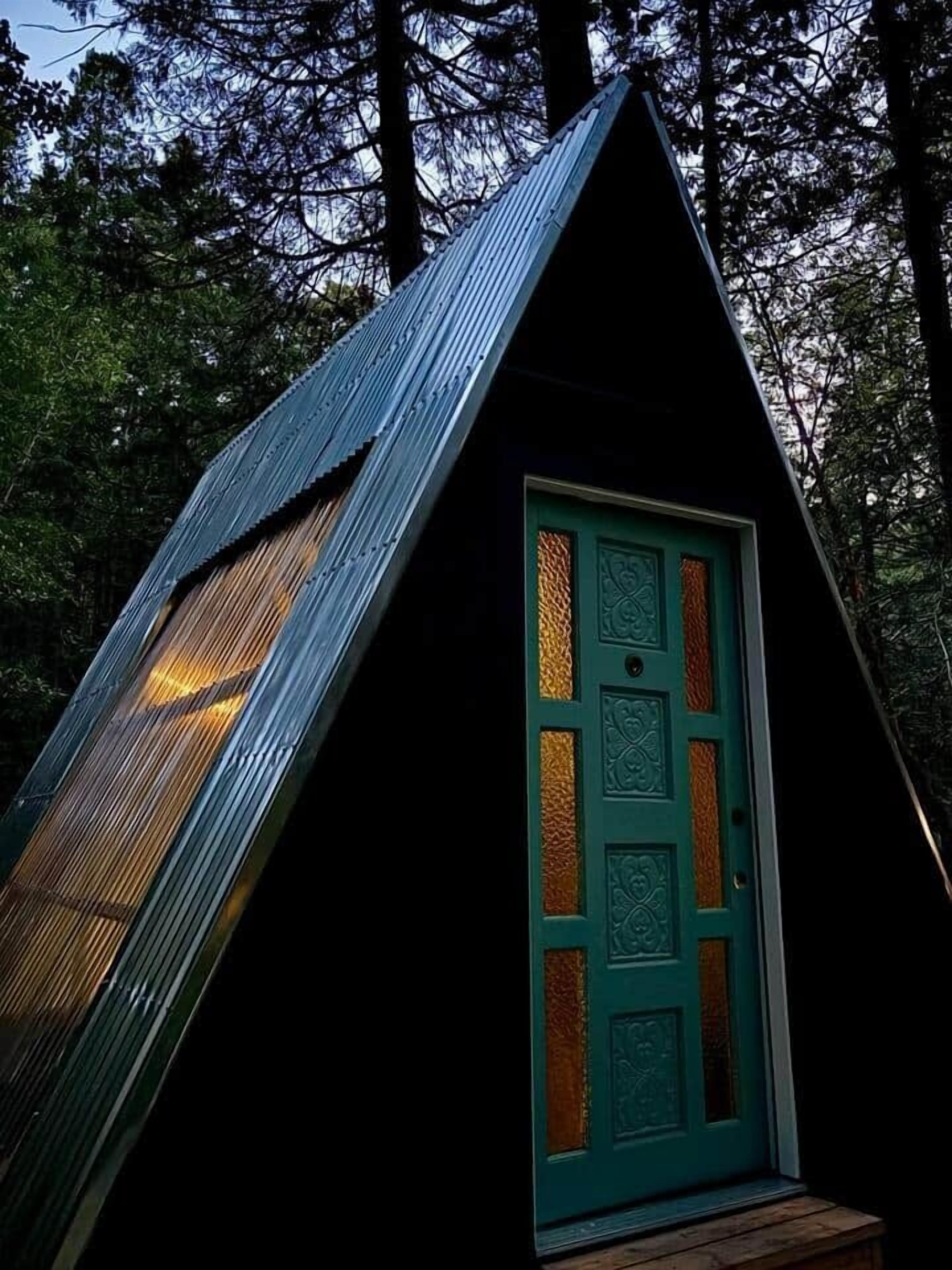 Una piccola baita di colore scuro con una porta verde e un tetto spiovente, circondata da alberi.