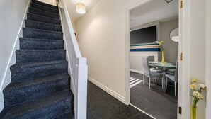A staircase with dark carpeted steps and white handrails, leading to a room with a striped wall and a table with chairs. - Six‑bedroom stay with garden, private hot tub, parking and easy Manchester tram links (Greater Manchester)