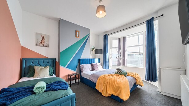 A modern bedroom with two beds, a large window with curtains, and a geometric wall design. - Six‑bedroom stay with garden, private hot tub, parking and easy Manchester tram links (Greater Manchester)