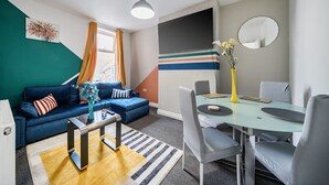 A modern living room with a blue sofa, a glass coffee table, and a dining area with a round table and chairs. - Six‑bedroom stay with garden, private hot tub, parking and easy Manchester tram links (Greater Manchester)