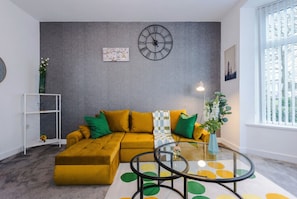 A living room with a sectional sofa, a glass coffee table, and a wall clock.
