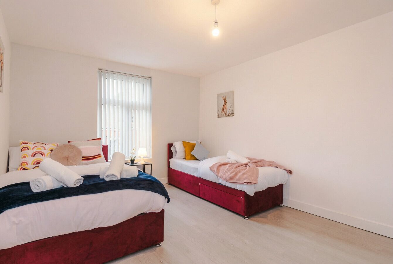 Ein Schlafzimmer mit zwei Betten, einem Fenster mit Jalousien, einem Wandbild und einem Nachttisch mit Lampe.