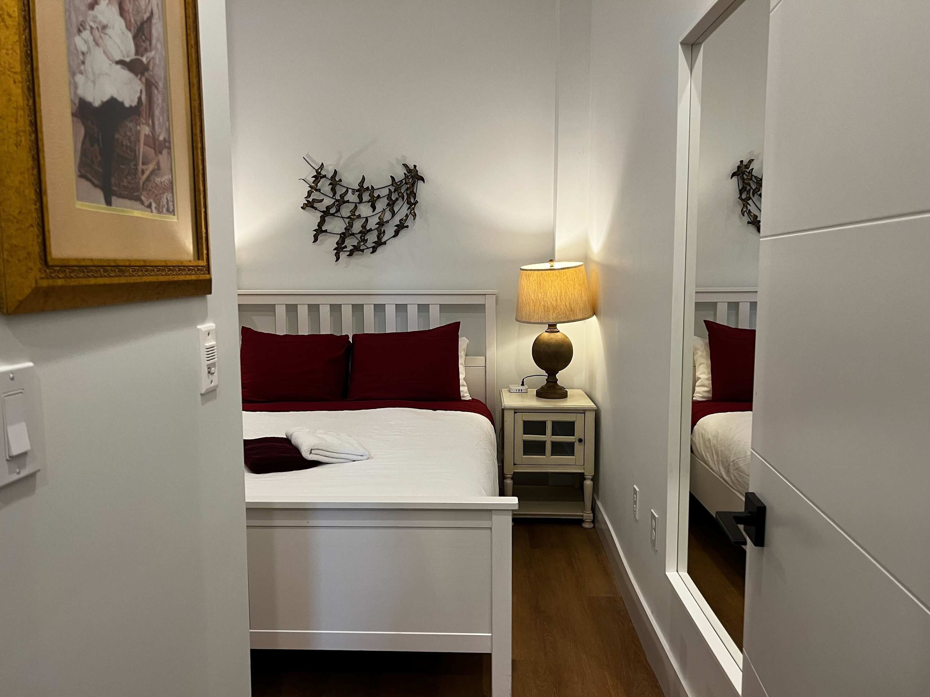 A hotel room with a bed, a bedside table with a lamp, a mirror, and a wall-mounted artwork.
