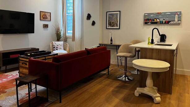 Een moderne woonkamer met een rode bank, een televisie, een eethoek met een kleine tafel en stoelen, en een keuken met een spoelbak en een decoratieve plank.