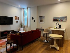 Een moderne woonkamer met een rode bank, een televisie, een eethoek met een kleine tafel en stoelen, en een keuken met een spoelbak en een decoratieve plank.