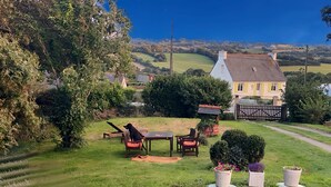 Ein sonniger Außenbereich mit Rasen, einem Tisch und Stühlen, umgeben von Topfpflanzen und Blick auf ein gelbes Haus im Hintergrund.
