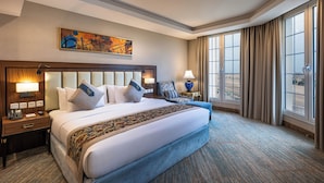 A hotel room with a large bed, a desk with a lamp, a chair, a painting on the wall, and a view through the window. - Braiden Hotel (Al Jubail)
