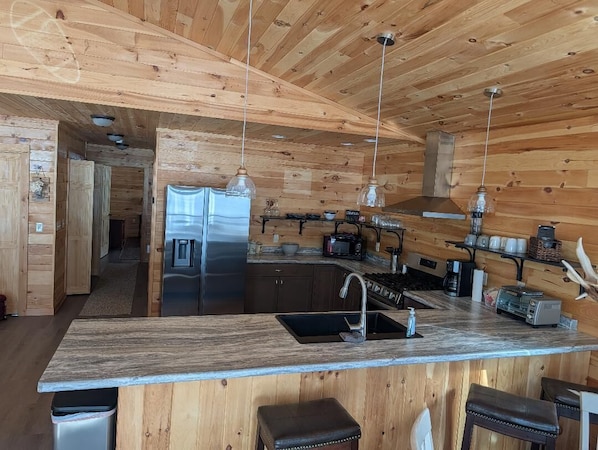 A wooden cabin interior with a kitchen area featuring a large island, stainless steel appliances, and hanging pendant lights.