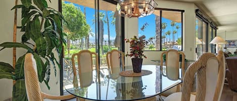 A dining area with a glass table, wicker chairs, and a view of palm trees through large windows.