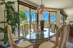A dining area with a glass table, wicker chairs, and a view of palm trees through large windows. - Exclusive 2BR, Golf Views & Private BBQ (Palm Desert)