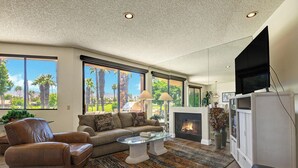 A living room with a fireplace, leather sofa, and a large window offering a view of palm trees. - Exclusive 2BR, Golf Views & Private BBQ (Palm Desert)
