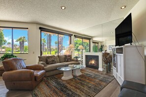 Ein Wohnzimmer mit Kamin, Ledersofa und großem Fenster mit Blick auf Palmen.