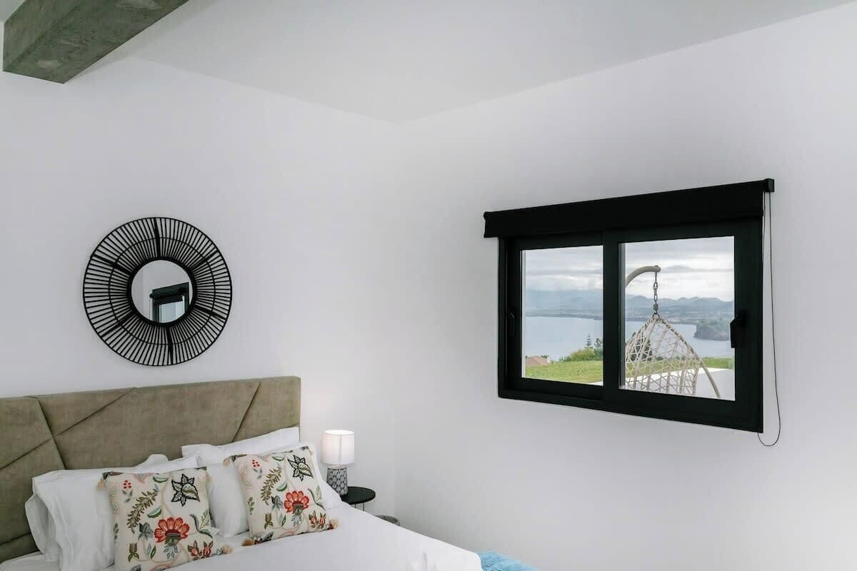 A bedroom with a bed, a circular wall decoration, a window with a view of a lake and mountains, and a nightstand with a lamp.