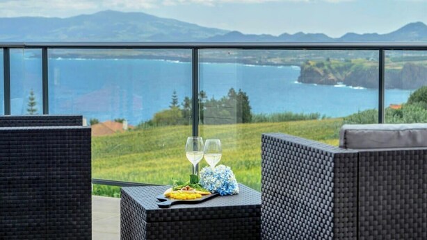 A balcony with wicker furniture, two glasses of white wine, and a plate of food. - GuestReady - Good Vibes | Sea Views, Terrace & BBQ (Ponta Delgada)