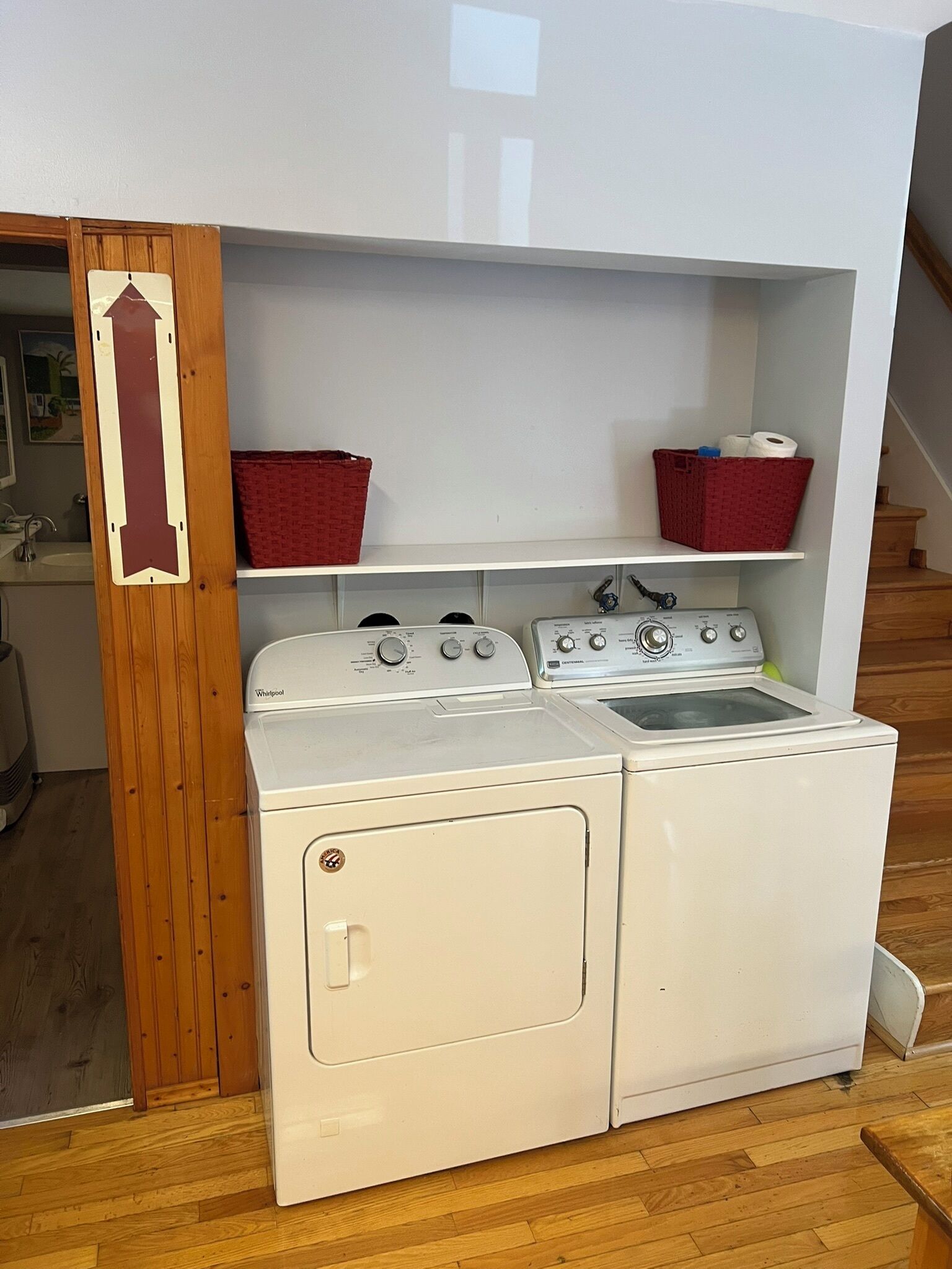 Een wasruimte met een voorlader wasmachine en droger, houten planken met manden, en een houten deur met een bordje.