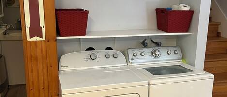 Een wasruimte met een voorlader wasmachine en droger, houten planken met manden, en een houten deur met een bordje.