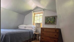 Une chambre avec un plafond en pente, un lit recouvert de draps blancs, une commode en bois, un petit tabouret et une fenêtre donnant sur un paysage verdoyant.